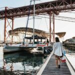 Un catamaran amarré dans une marina de Lisbonne sous le tablier rouge du pont du 25-Avril, au soleil couchant