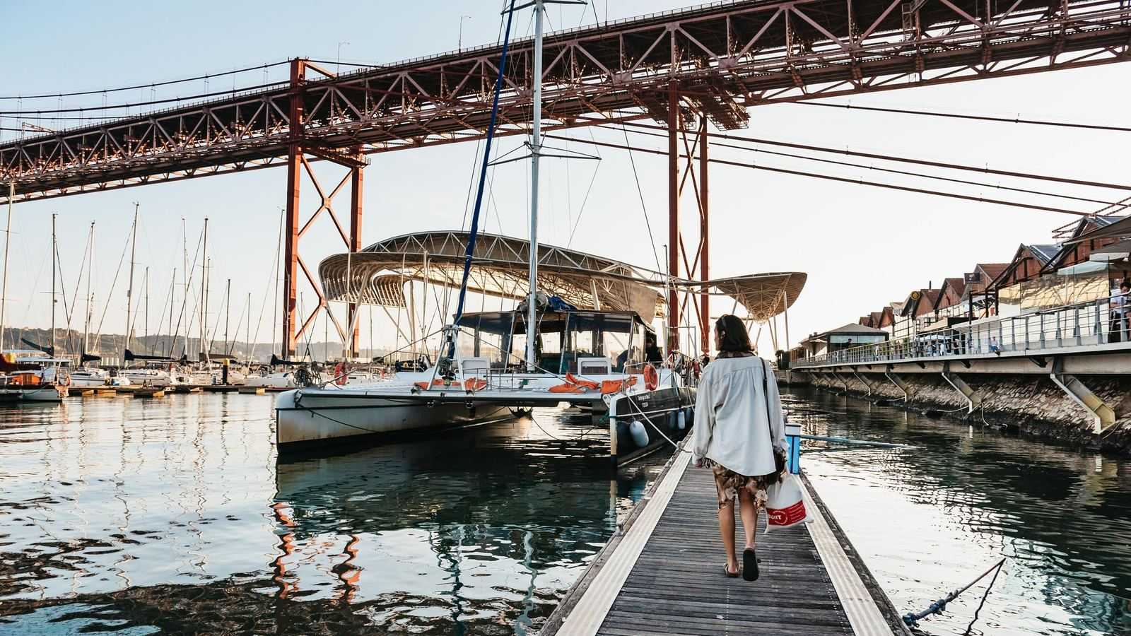 Un catamaran amarré dans une marina de Lisbonne sous le tablier rouge du pont du 25-Avril, au soleil couchant