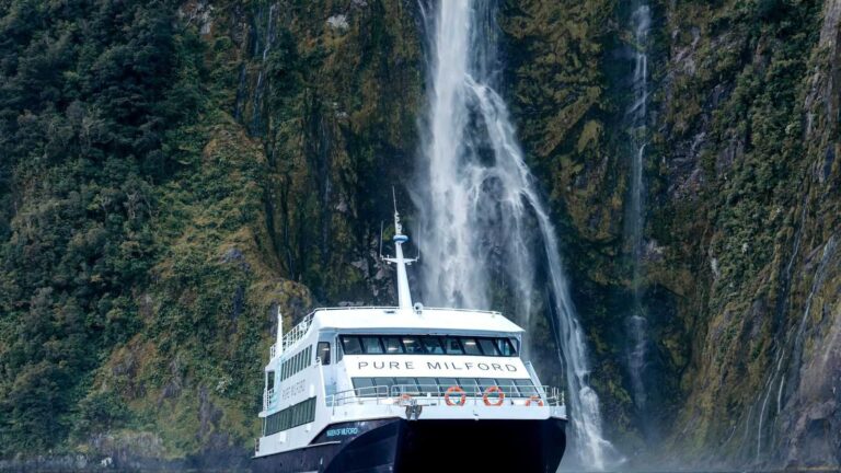 Un bateau de croisière passe devant une cascade de Milford Sound, encaissée entre les parois du fjord
